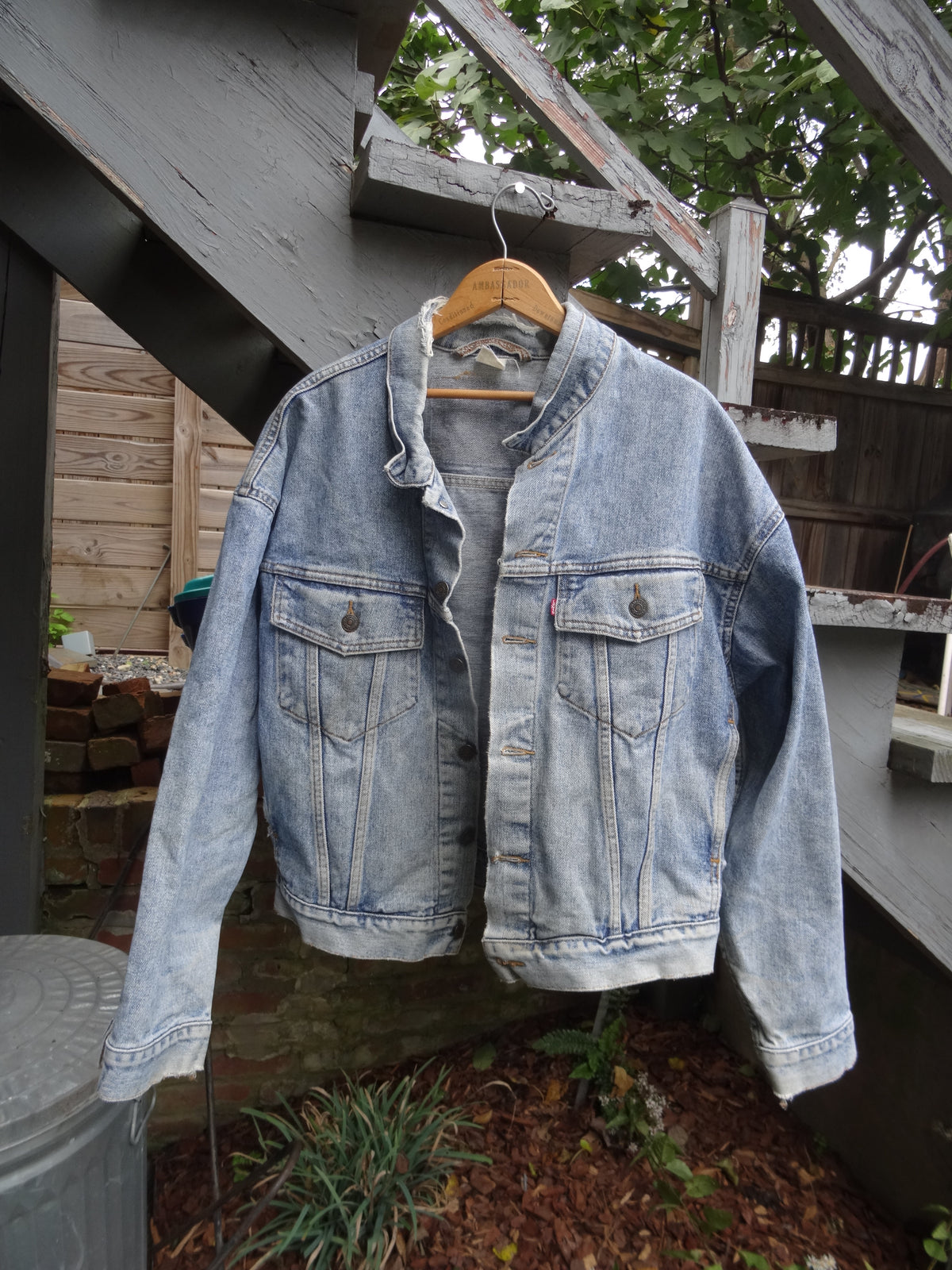 A light-wash blue denim jacket hanging on a wooden hanger outdoors on a porch or deck, with visible wood beams, fence, plants, and mulch in the background.